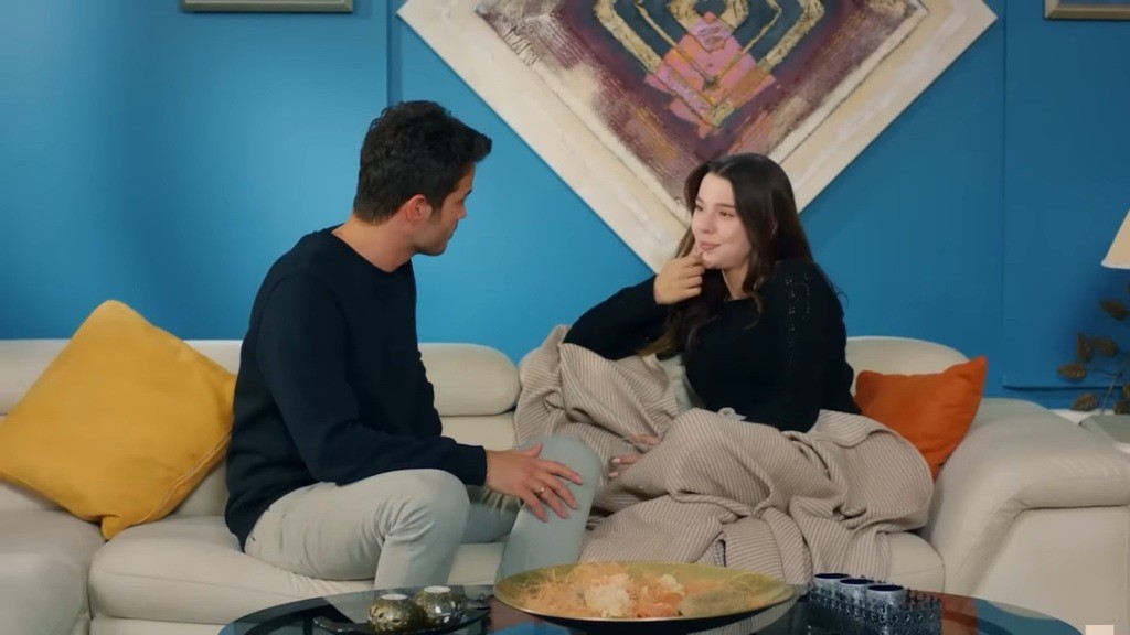 Zeynep siedzi na sofie, owinięta w koc. Obok niej znajduje się Ege.
