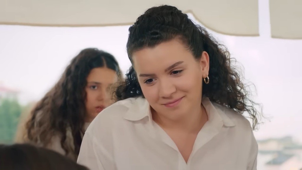 Melis uśmiecha się lekko, patrząc na Zeynep. Za jej plecami widać Melodi.