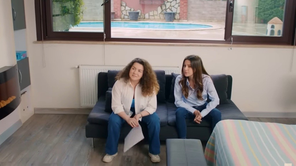 Melis i Zeynep siedzą na sofie. Melis trzyma w rękach dokument.