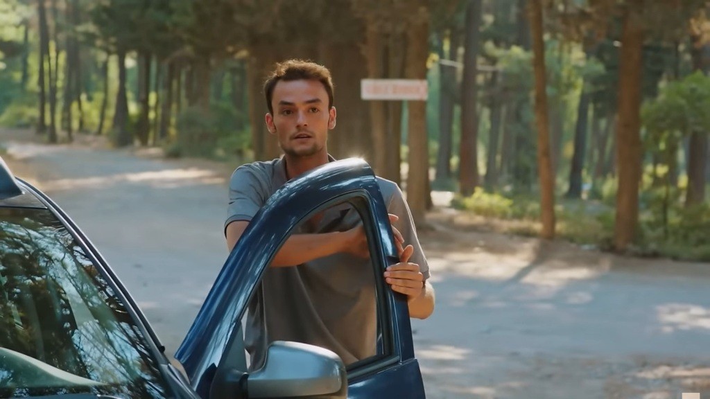 Alper stoi przy otwartych drzwiach samochodu.
