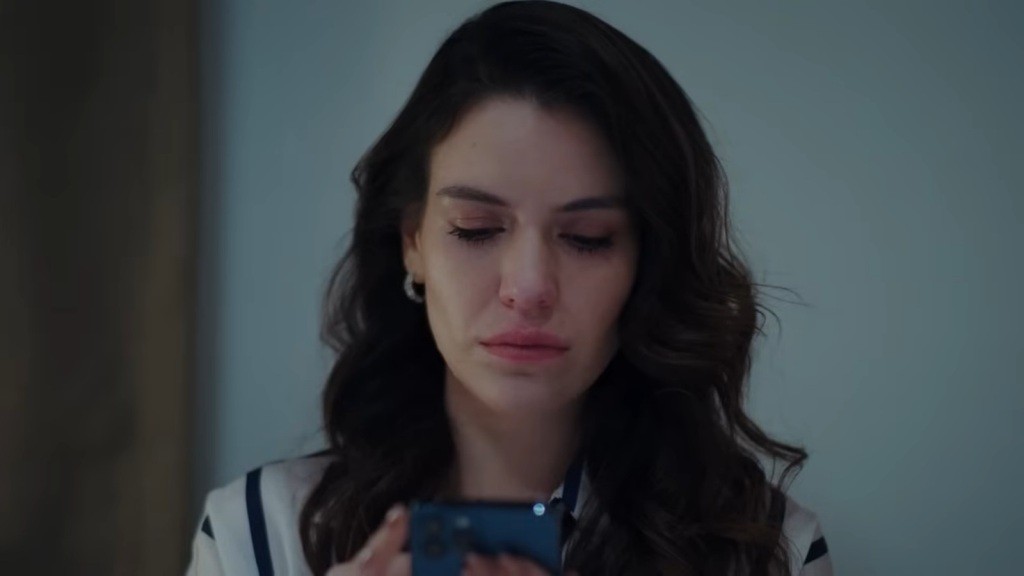 Pelin trzyma telefon w rękach i wpatruje się w jego ekran. Jej twarz wyraża głęboki smutek.