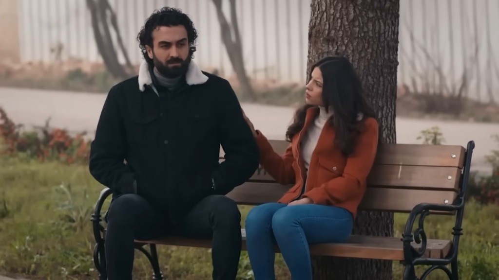 Ferit i Ayse siedzą na ławce w parku. Ona trzyma dłoń na jego ramieniu.