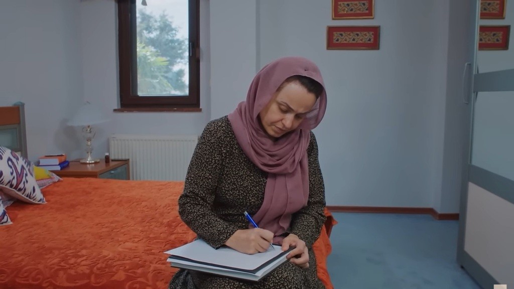 Cavidan siedzi na skraju łóżku, pisząc w notatniku.