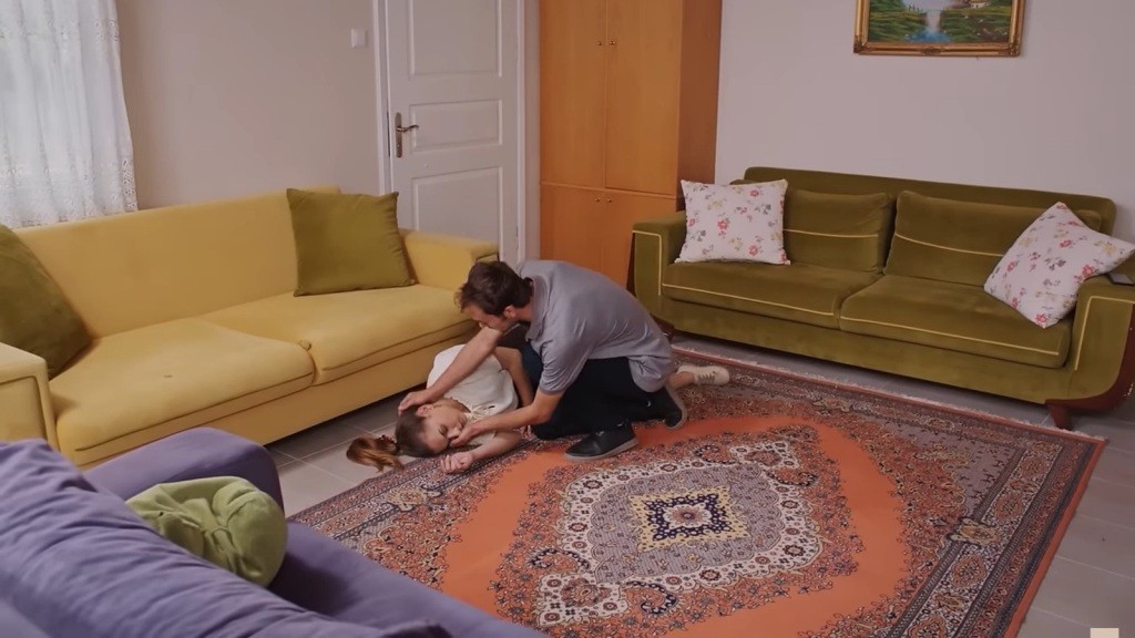 Alper pochyla się nad nieprzytomną Silą, która upadła obok kanapy.