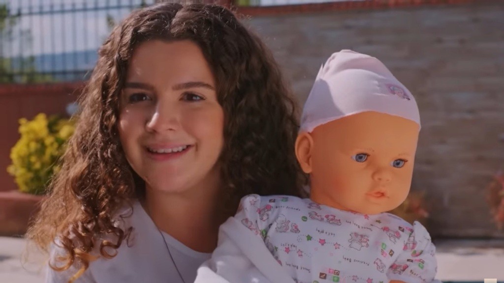 Melis uśmiecha się, trzymając w ramionach lalkę niemowlę.