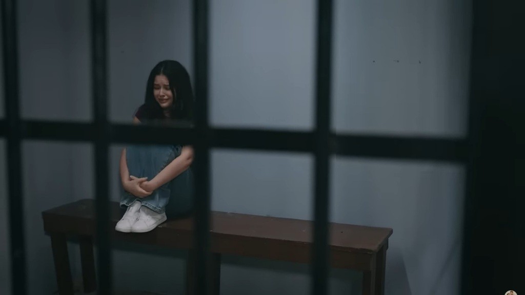 Merve siedzi na ławce w celi, przyciągając kolana do brody.