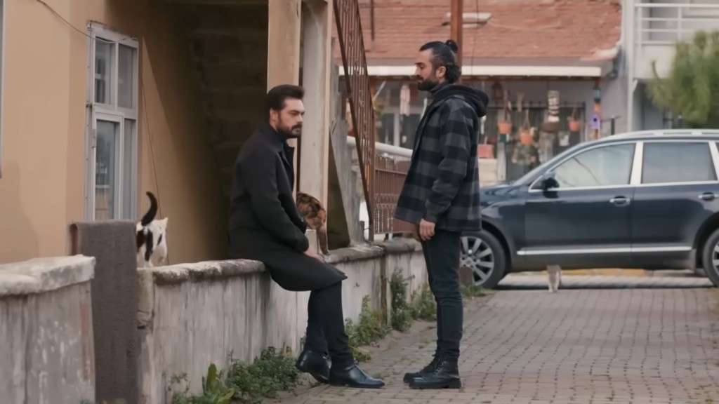 Yaman siedzi na murku, po którym chodzi kot. Naprzeciwko niego stoi Ferit. Znajdują się w biednej dzielnicy.