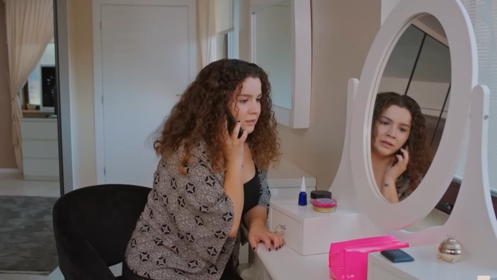Melis siedzi przy toaletce i rozmawia przez telefon.