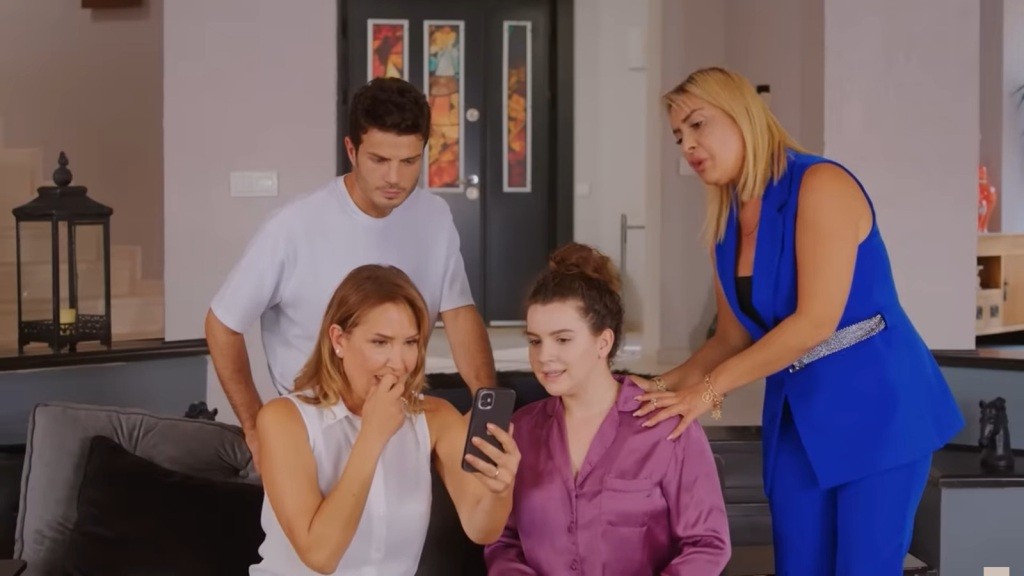 Feraye i Melis siedzą na sofie, a tuż za nimi stoją Ege i Belkis. Wszyscy wpatrują się w telefon, który trzyma Feraye. Ich twarze wyrażają ciekawość i napięcie.