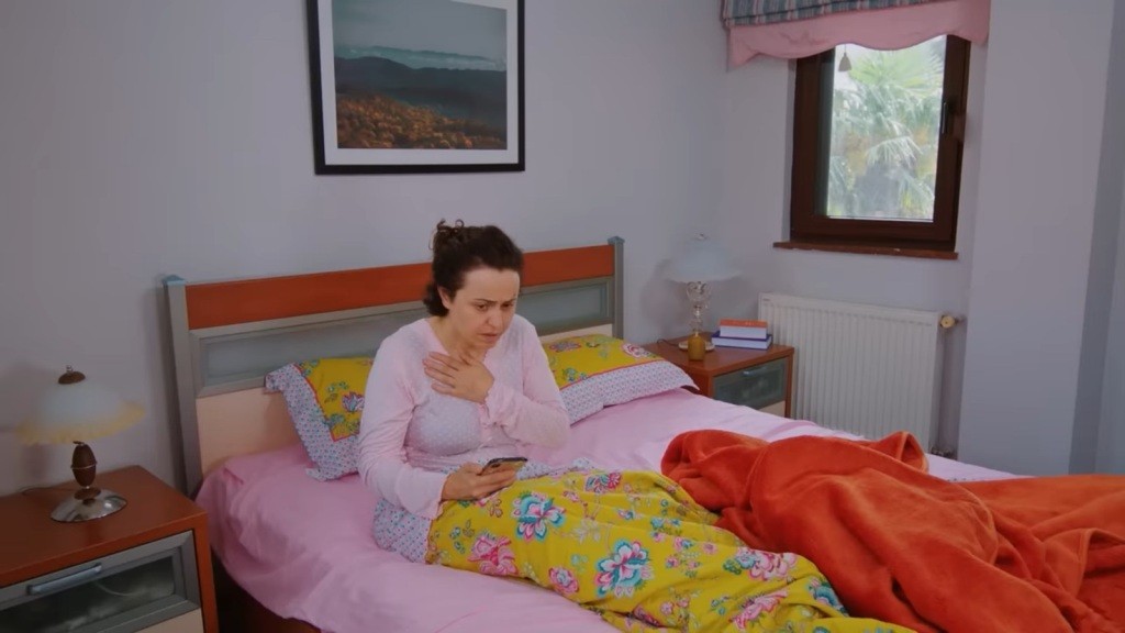 Cavidan leży na łóżku, trzymając telefon w jednej ręce, a drugą przyciskając do piersi, jakby w geście przerażenia.