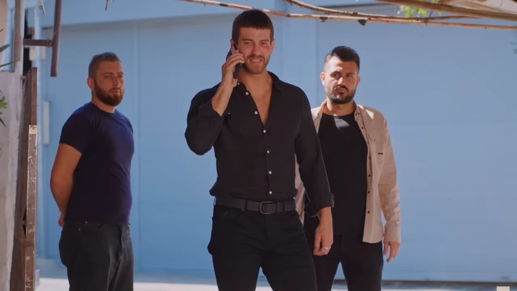 Cihan rozmawia przez telefon. Za nim stoi dwóch mężczyzn.