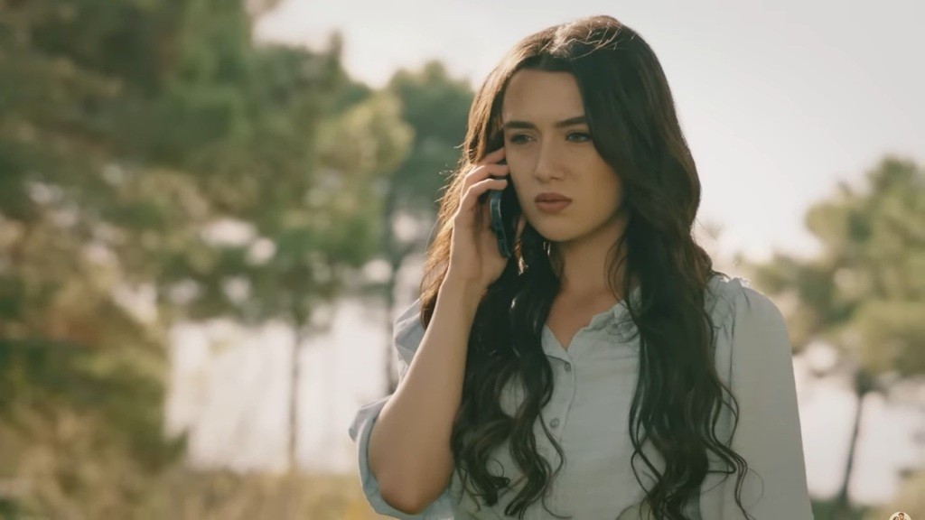 Zeynep rozmawia przez telefon, a za nią widać korony drzew, rozświetlone promieniami słońca, które tworzą malowniczy, złocisty pejzaż.