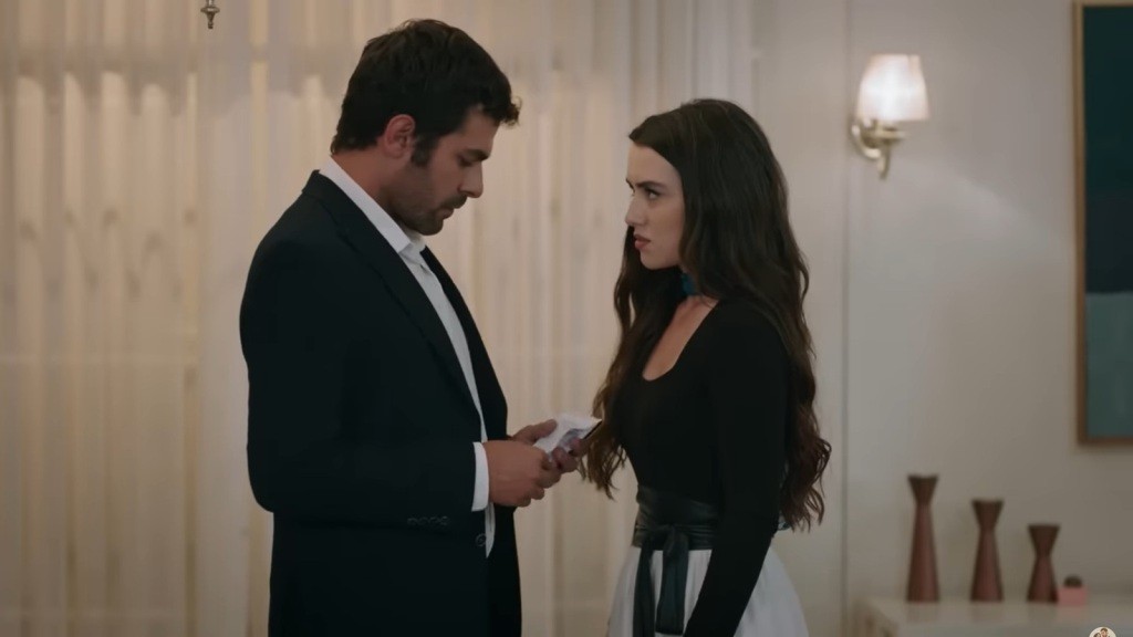 Halil stoi przed Zeynep. W rękach trzyma książeczkę czekową.