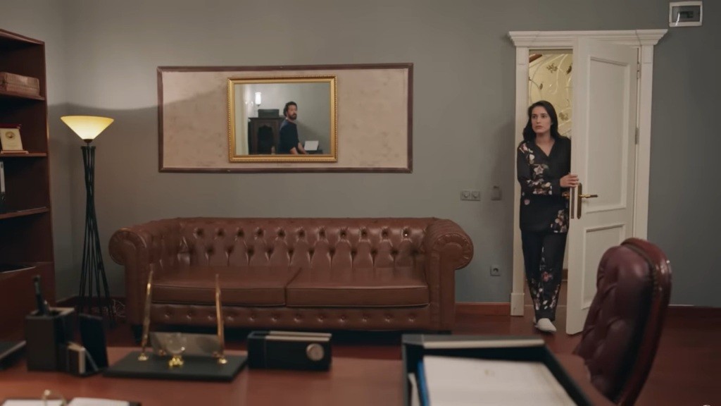 Gulhan zagląda do gabinetu. W lustrze widać odbicie jej męża.