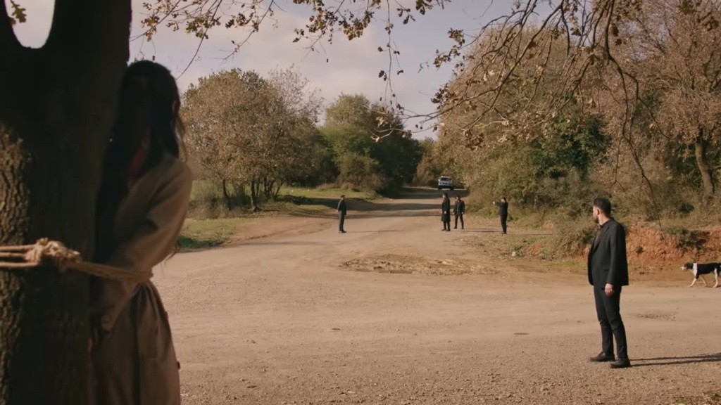 Zeynep jest przywiązana do drzewa. Na drodze widać stojących mężczyzn.