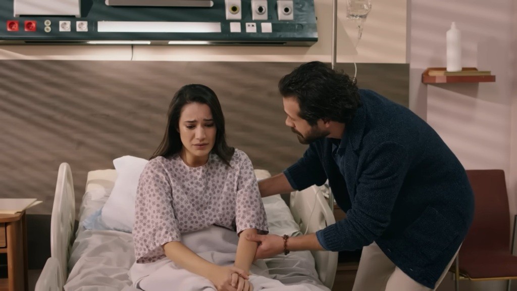 Gulhan siedzi na szpitalnym łóżku. Tekin pochyla się nad nią, delikatnie ujmując jej ramię.