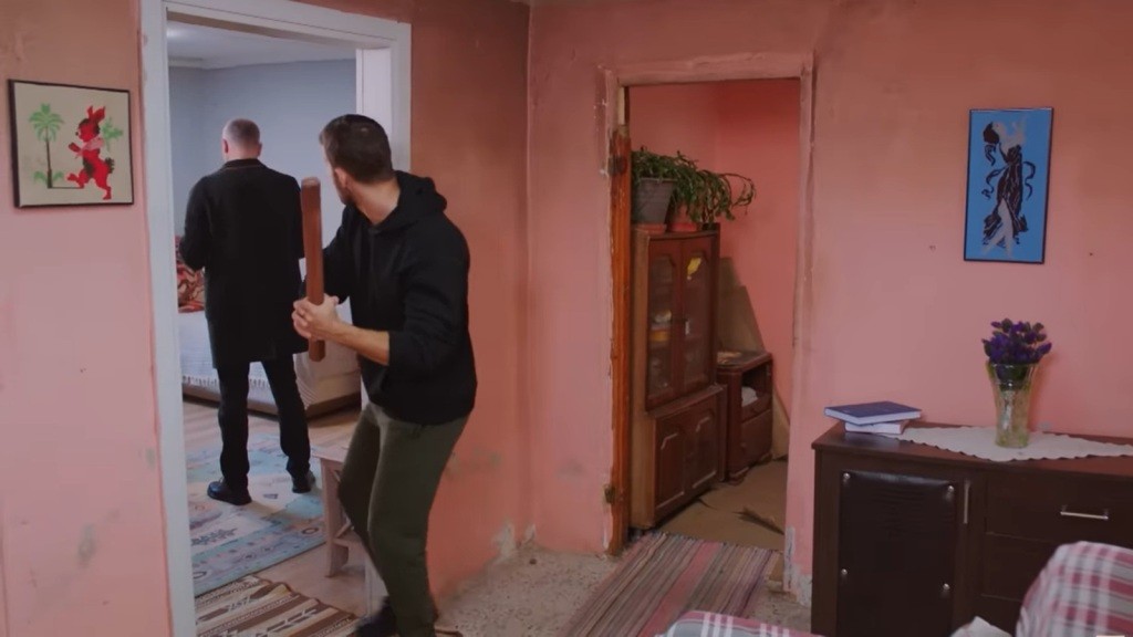 Cihan zbliża się ostrożnie, trzymając w ręku polano, kierując się w stronę pokoju, w którym Bulent stoi odwrócony plecami.