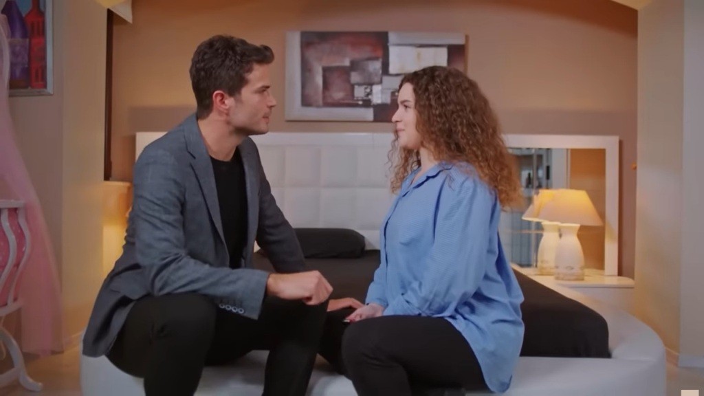 Ege i Melis siedzą na skraju łóżka, zwróceni do siebie twarzami.