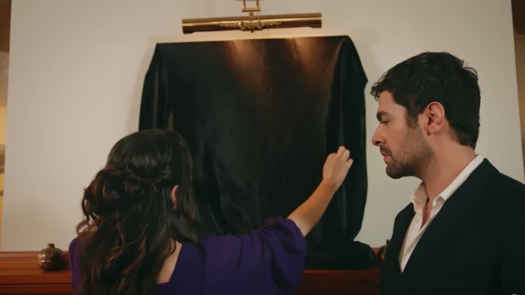 Zeynep zdejmuje płótno, którym zasłonięty jest obraz na ścianie.