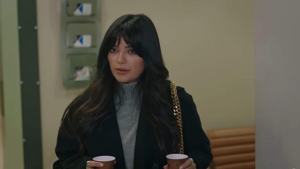 Gozde stoi w szpitalnym korytarzu, trzymając dwa kubki w rękach.