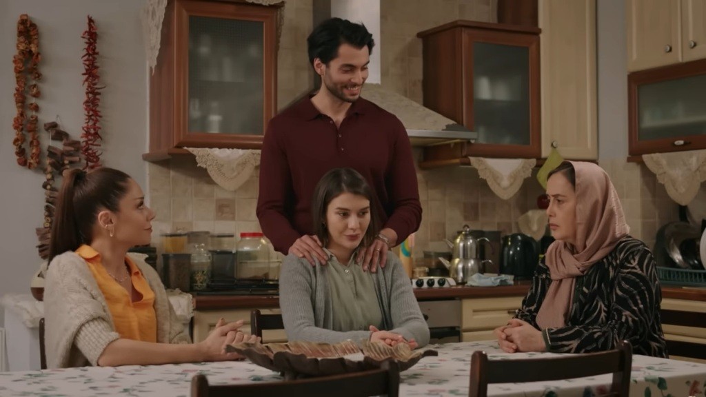 Kumru, Selma i Kiymet siedzą przy stole w kuchni. Eren trzyma ręce na ramionach żony, stojąc tuż za nią.