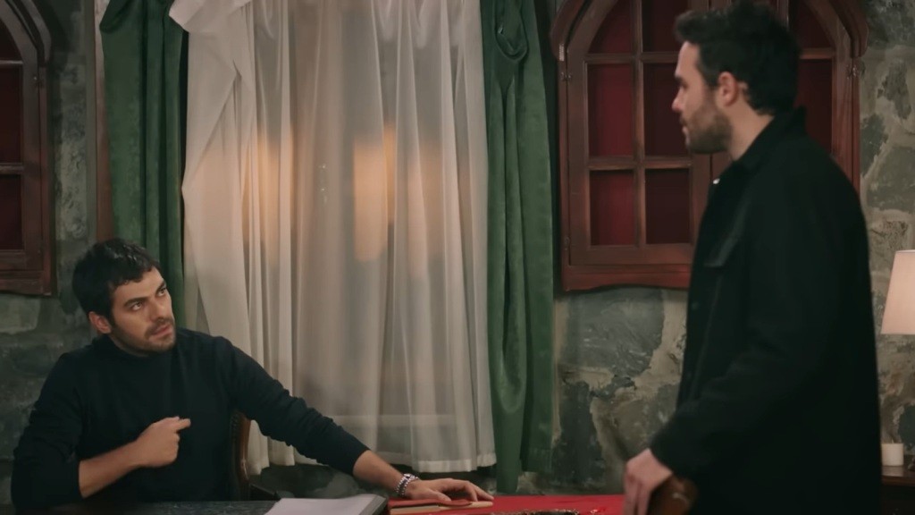 Halil siedzi za biurkiem w pracowni. Pokazuje palcem na swoje serce, rozmawiając z Hakanem.