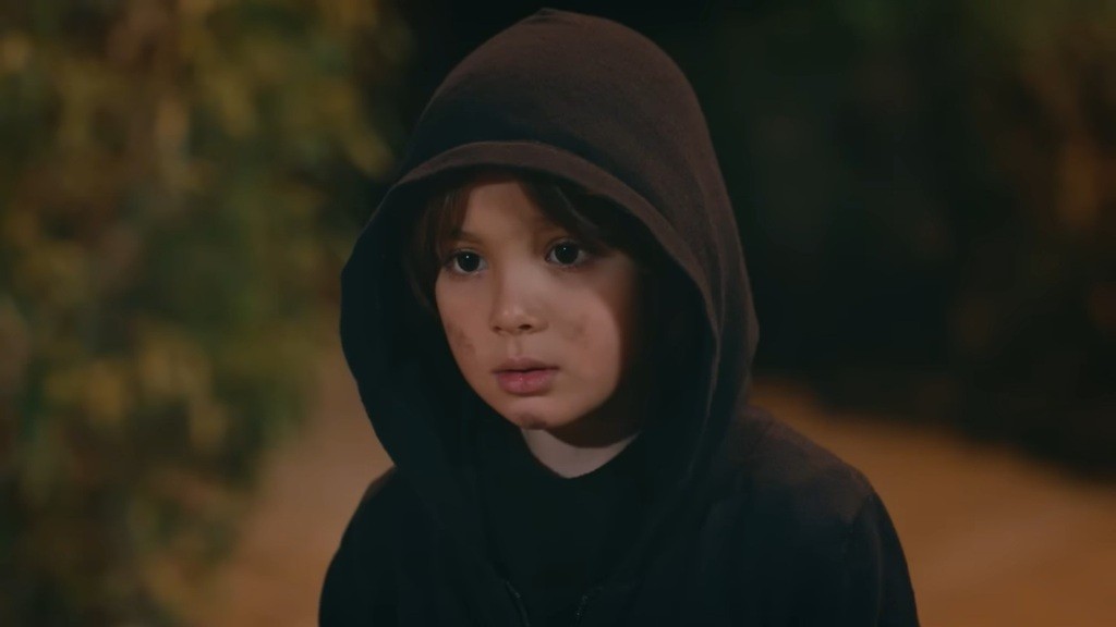 Zbliżenie na twarz chłopca, na której widać strach. Ma pobrudzone policzki i kaptur naciągnięty głęboko na głowę.