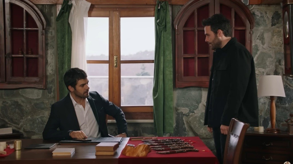 Halil siedzi przy biurku w pracowni. Obok stoi Hakan.