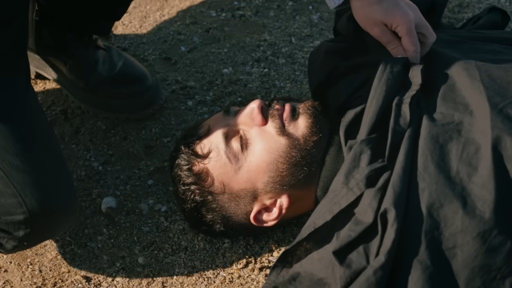 Policjant podnosi materiał, odsłaniając twarz znalezionego na plaży denata.