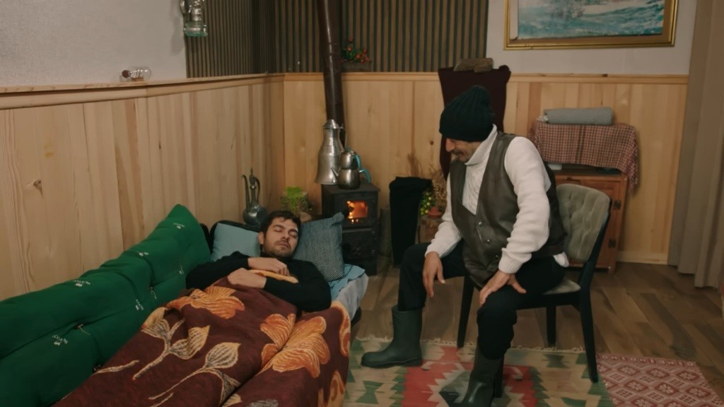 Halil śpi na łóżku, a Yunus siedzi obok na krześle.