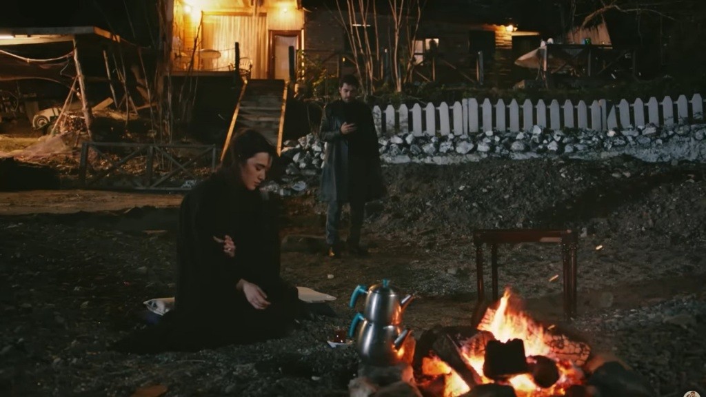 Zeynep siedzi przy ognisku. Halil stoi nieco dalej, z telefonem w ręku.