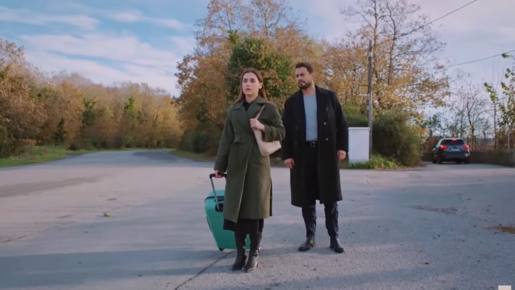 Sila idzie drogą, ciągnąc za sobą walizkę. Za nią stoi Kuzey.