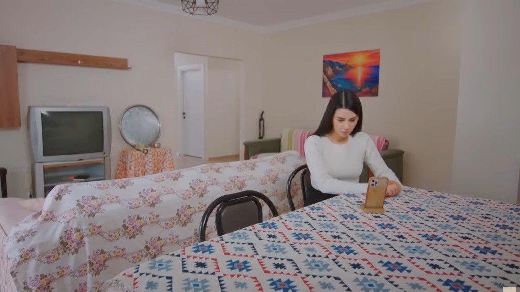 Bahar siedzi przy stole w skromnym mieszkaniu. Przed nią na podstawce stoi telefon.