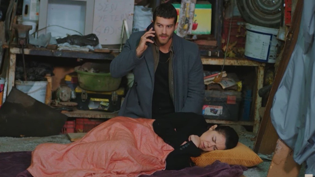 Cihan klęczy obok Gonul, która śpi na posłaniu w garażu. Z telefonem przy uchu prowadzi cichą rozmowę.