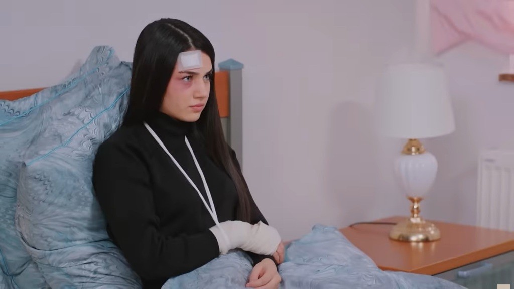 Bahar leży na łóżku, oparta plecami o wezgłowie. Na jej czole widnieje biały plaster, a ramię jest unieruchomione w temblaku.