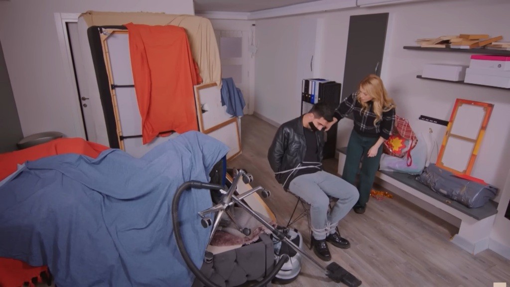 Gokhan jest przywiązany do krzesła w zagraconym pomieszczeniu. Obok stoi Seda.