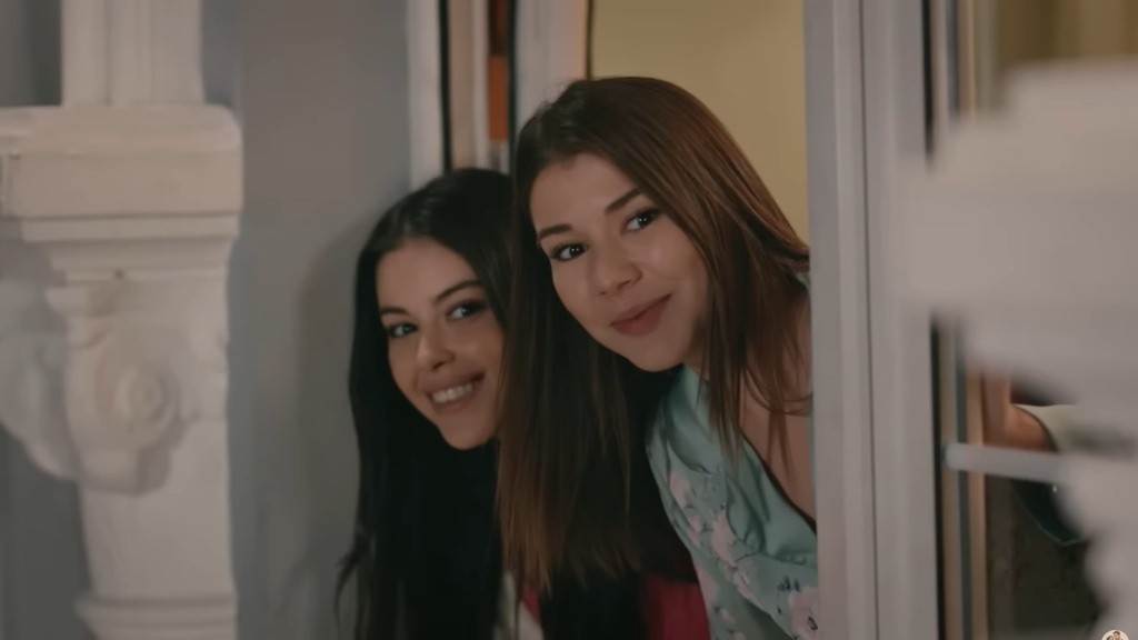 Merve i Selma wyglądają przez drzwi balkonowe. Ich twarze są pełne radości.