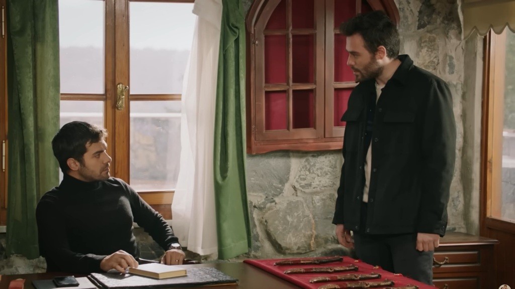 Halil siedzi przy biurku. Obok stoi Hakan.