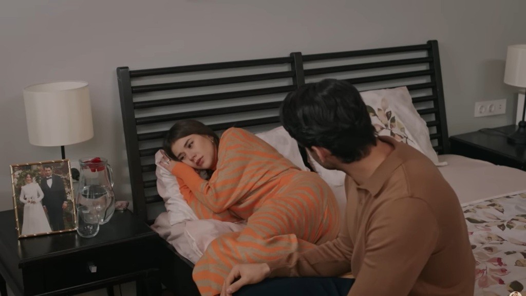 Selma leży na łóżku, przytulając policzek do poduszki. Obok niej siedzi Eren.