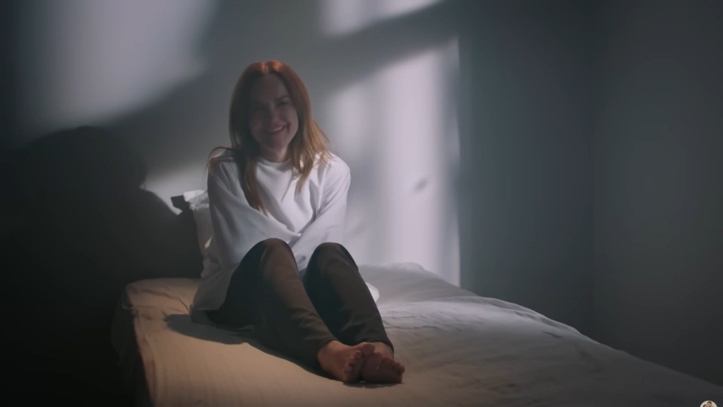 Songul siedzi na łóżku w kaftanie bezpieczeństwa. Uśmiecha się szaleńczo.