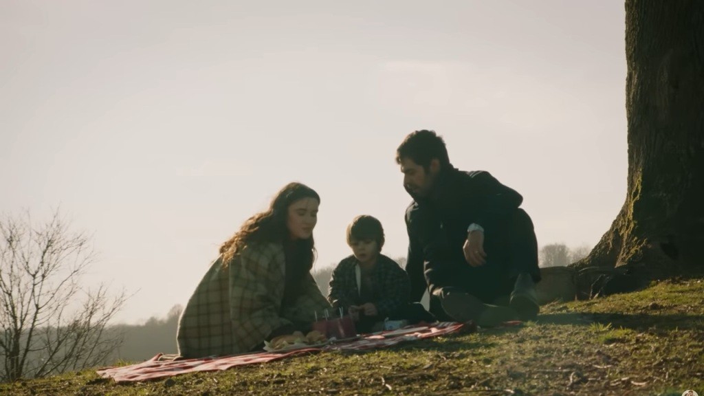 Halil, Zeynep i ich syn siedzą na kocu pod drzewem na wzgórzu.