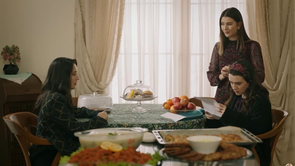 Zeynep i Gulhan siedzą przy stoliku, w milczeniu przepełnionym oczekiwaniem. Gulhan nerwowo zerka w stronę Zeynep, która z uwagą studiuje list. Obok nich Selma stoi, zacierając ręce z ekscytacją, pełna nadziei na to, co ten moment może przynieść.