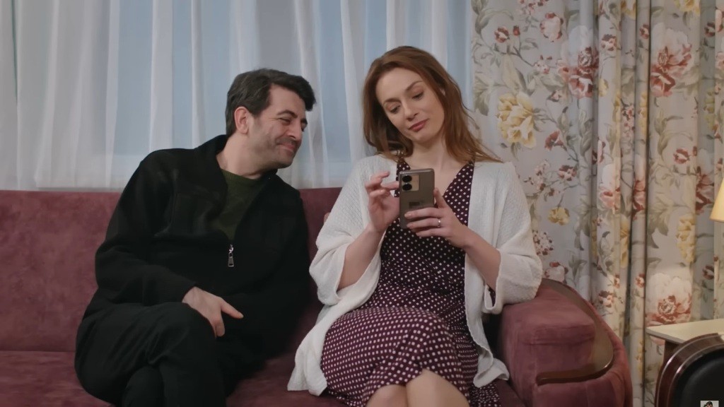 Cemil i Derya siedzą na kanapie. Mężczyzna patrzy na trzymane w jej rękach telefon.