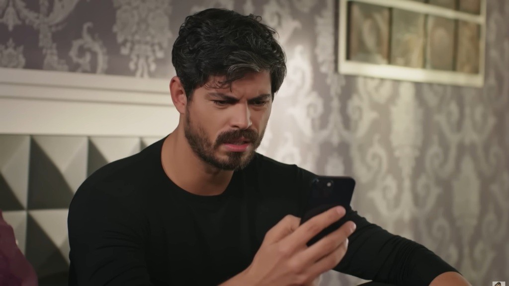Cihan wpatruje się w ekran telefonu, marszcząc brwi. Na jego twarzy zaskoczenie miesza się ze złością.