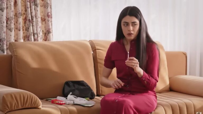 Hancer siedzi na kanapie. W ręku trzyma strzykawkę. Obok niej leżą liczne opakowania z lekarstwami.