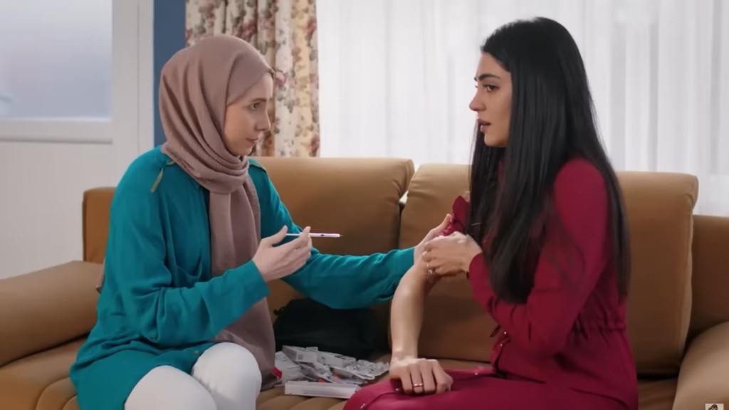 Hancer podwija rękaw bluzki, a siedząca obok Sinem unosi strzykawkę, by zrobić jej zastrzyk.