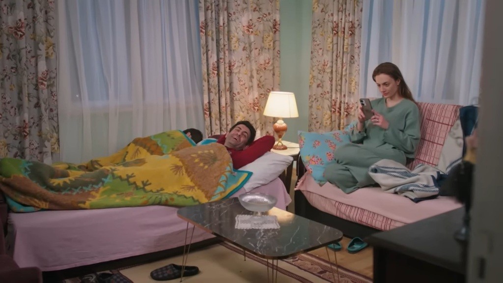 Cemil leży na rozłożonej wersalce, przykryty kocem. Na drugiej wersalce siedzi Derya, wpatrzona w ekran telefonu.