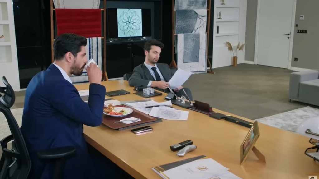 Cihan pije kawę i je śniadanie, siedząc przy biurku w firmie. Po drugiej stronie biurka siedzi Engin i zapoznaje się ze szkicami.