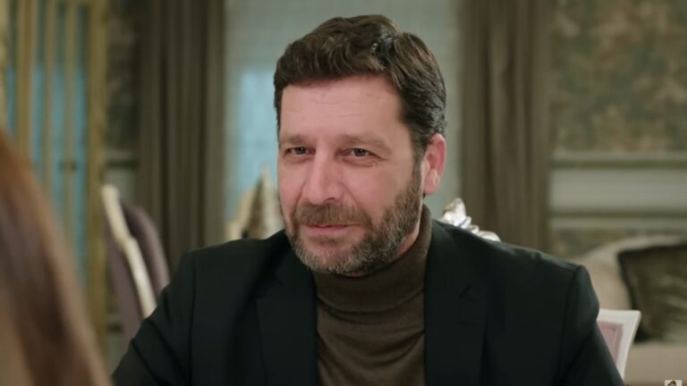 Zbliżenie na twarz Nusreta, siedzącego przy stole w salonie rezydencji Develioglu. Sprawia wrażenie zadowolonego.