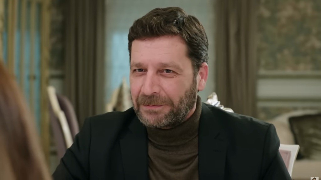 Zbliżenie na twarz Nusreta, siedzącego przy stole w salonie rezydencji Develioglu. Sprawia wrażenie zadowolonego.